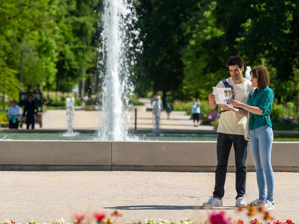BadLippspringe-Landesgartenschau-Teutoburger-Wald-Tourismus-Patrick-Gawandtka-034.jpg Junges Paar betrachtet eine Karte im Park vor einem beeindruckenden Springbrunnen im Hintergrund.