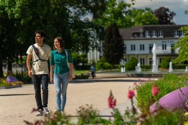 BadLippspringe-Landesgartenschau-Teutoburger-Wald-Tourismus-Patrick-Gawandtka-041.jpg Ein Paar spaziert im Sommer lächelnd durch einen blühenden Park mit einem Hotel im Hintergrund.