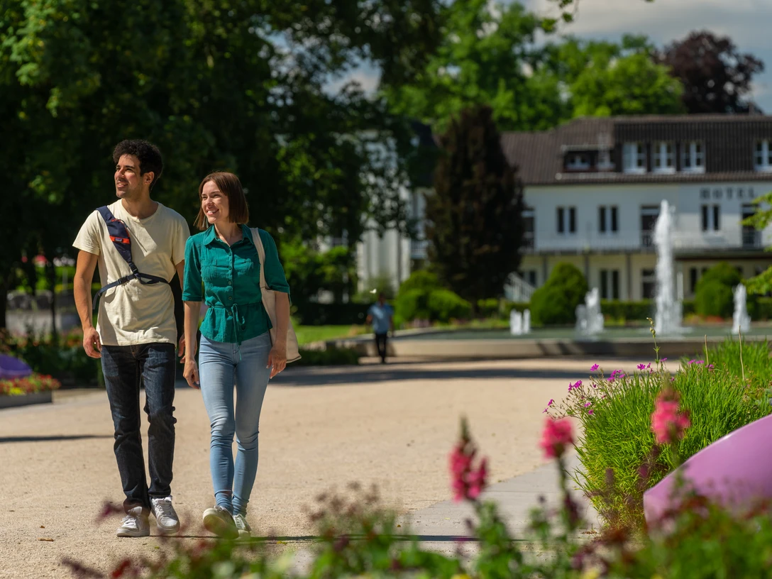 BadLippspringe-Landesgartenschau-Teutoburger-Wald-Tourismus-Patrick-Gawandtka-041.jpg Ein Paar spaziert im Sommer lächelnd durch einen blühenden Park mit einem Hotel im Hintergrund.