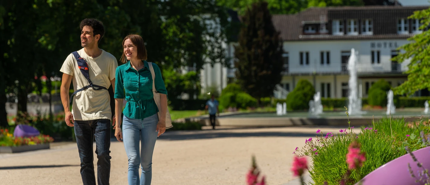 BadLippspringe-Landesgartenschau-Teutoburger-Wald-Tourismus-Patrick-Gawandtka-041.jpg Ein Paar spaziert im Sommer lächelnd durch einen blühenden Park mit einem Hotel im Hintergrund.