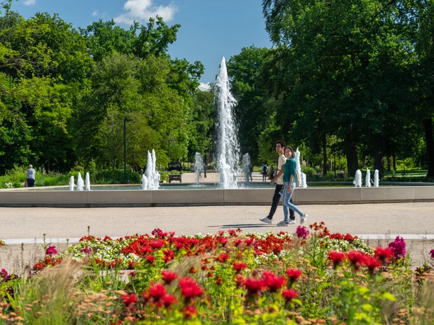 BadLippspringe-Landesgartenschau-Teutoburger-Wald-Tourismus-Patrick-Gawandtka-032.jpg Zwei Personen spazieren an einem blühenden Blumenbeet vor einem großen Springbrunnen im Park vorbei.