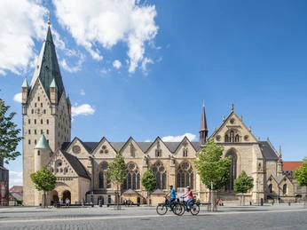 Paderborn-Dom-Teutoburger-Wald-Tourismus-Patrick-Gawandtka-073.jpg Paderborner Dom mit markantem Turm, umgeben von Bäumen, Radfahrer im Vordergrund bei Sonnenschein.