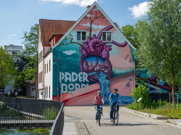 Paderborn-Herzgraffiti-Teutoburger-Wald-Tourismus-Patrick-Gawandtka-078.jpg Fahrradfahrer auf sonnigem Weg vor farbenfrohem Haus mit kunstvollem Wandgemälde in Paderborn.