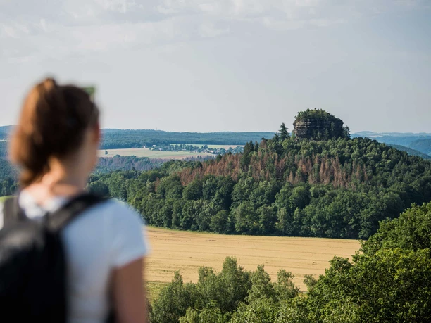 Uitzicht vanaf de Kaiserkrone naar de Zirkelstein Een vrouw met rugzak kijkt uit over een beboste heuvel met rotsen in een groen landschap onder een blauwe lucht.