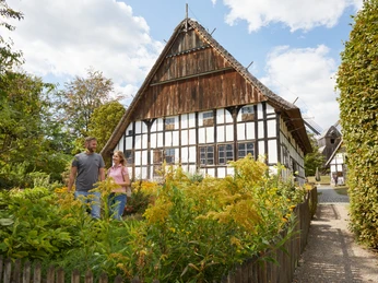Bielefeld-Bauernhausmuseum-Teutoburger-Wald-Tourismus-T-Evers-057.jpg Ein Paar spaziert vor einem historischen, mit Fachwerk versehenen Gebäude bei sonnigem Wetter entlang.