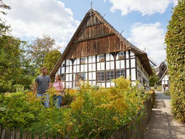 Bielefeld-Bauernhausmuseum-Teutoburger-Wald-Tourismus-T-Evers-057.jpg Ein Paar spaziert vor einem historischen, mit Fachwerk versehenen Gebäude bei sonnigem Wetter entlang.