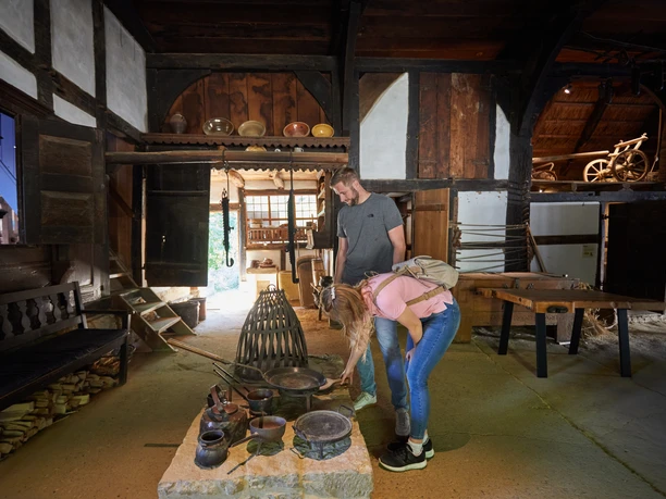 Zwei Personen erkunden ein historisches Bauernhausmuseum, umgeben von alten Küchenutensilien.