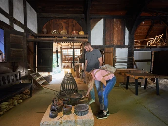 Bielefeld-Bauernhausmuseum-Teutoburger-Wald-Tourismus-T-Evers-044.jpg Zwei Personen erkunden ein historisches Bauernhausmuseum, umgeben von alten Küchenutensilien.