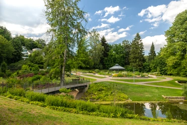 Bielefeld-Botanischer Garten-Teutoburger-Wald-Tourismus-D-Ketz-095.jpg Ein idyllischer Park in Bielefeld mit Teich, Brücke, Blumenbeeten und einem Pavillon unter blauem Himmel.