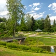 Bielefeld-Botanischer Garten-Teutoburger-Wald-Tourismus-D-Ketz-095.jpg Ein idyllischer Park in Bielefeld mit Teich, Brücke, Blumenbeeten und einem Pavillon unter blauem Himmel.