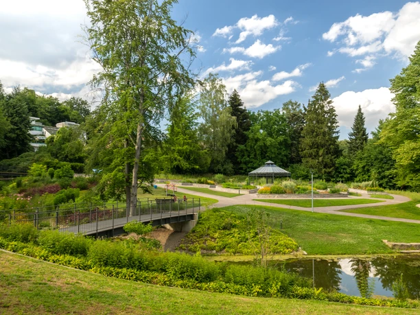 Bielefeld-Botanischer Garten-Teutoburger-Wald-Tourismus-D-Ketz-095.jpg Ein idyllischer Park in Bielefeld mit Teich, Brücke, Blumenbeeten und einem Pavillon unter blauem Himmel.