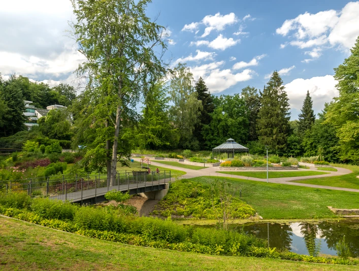 Bielefeld-Botanischer Garten-Teutoburger-Wald-Tourismus-D-Ketz-095.jpg Ein idyllischer Park in Bielefeld mit Teich, Brücke, Blumenbeeten und einem Pavillon unter blauem Himmel.
