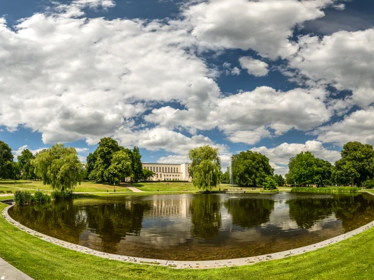 Ein idyllischer Park mit einem Teich in der Mitte, umgeben von grünen Bäumen und Rasenflächen unter einem wolkigen Himmel.