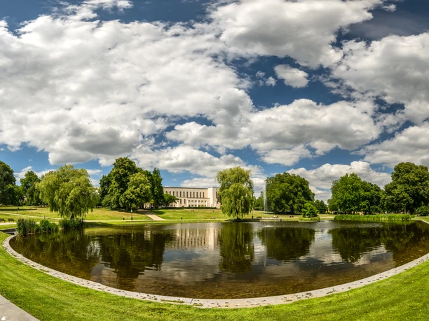 Ein idyllischer Park mit einem Teich in der Mitte, umgeben von grünen Bäumen und Rasenflächen unter einem wolkigen Himmel.