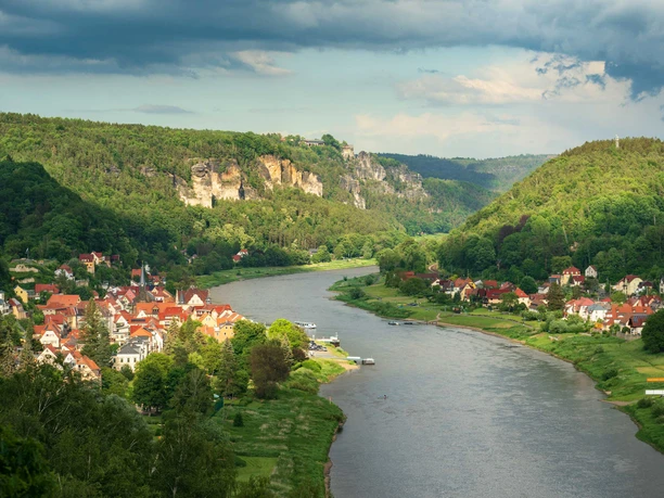 Pohled z Wilkeaussicht Říční krajina s vesnicí s červenými střechami na břehu, obklopená zelenými kopci a zalesněnými útesy pod zataženou oblohou.