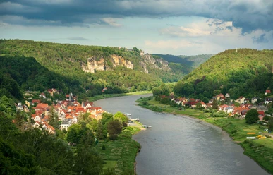View from the Wilkeaussicht River landscape with a village with red roofs on the bank, surrounded by green hills and wooded cliffs under a cloudy sky.
