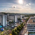 Panorama des Universitätsgeländes Bielefeld mit Gebäuden, Menschenmengen und sonnigem Himmel.