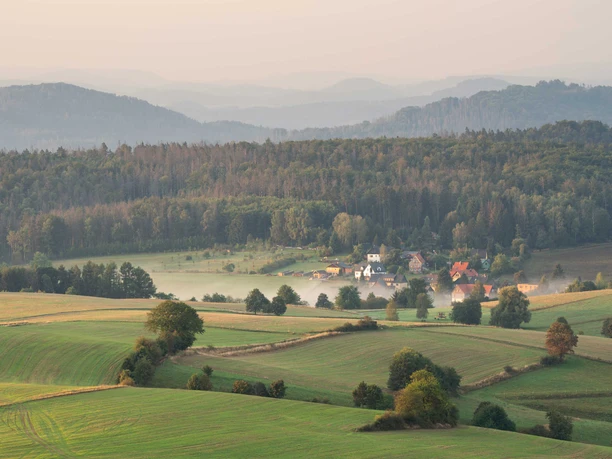 View from the Weifberg Tower Green hills with scattered trees, a small village in the valley and a misty atmosphere, surrounded by dense forests and gentle mountains in the background.