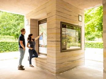 Entdeckungsfreudige betrachten eine Informationskarte in einem überdachten Bereich mit grüner Umgebung.