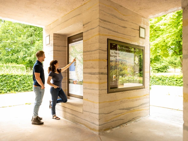 Bielefeld-Johannisberg-Teutoburger-Wald-Tourismus-D-Ketz-060.jpg Entdeckungsfreudige betrachten eine Informationskarte in einem überdachten Bereich mit grüner Umgebung.