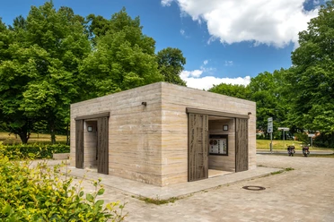 Öffentliche Toilettenanlage in einem Park, umgeben von Bäumen und grünem Laubwerk.