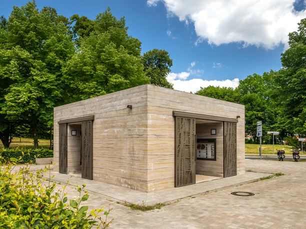 Öffentliche Toilettenanlage in einem Park, umgeben von Bäumen und grünem Laubwerk.