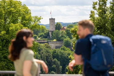 Ein Paar genießt den Blick auf eine Burgruine, umgeben von üppigem Grün bei sonnigem Wetter.