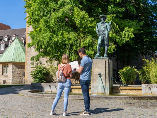 <p>Ein Paar betrachtet eine Karte vor einer Statue in einem sonnigen, grünen Platz mit Brunnen.</p>