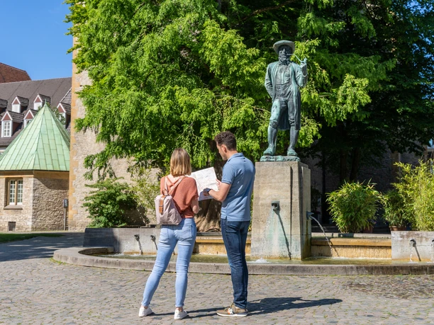 Bielefeld-Kirchplatz-Teutoburger-Wald-Tourismus-Patrick-Gawandtka-011.jpg <p>Ein Paar betrachtet eine Karte vor einer Statue in einem sonnigen, grünen Platz mit Brunnen.</p>