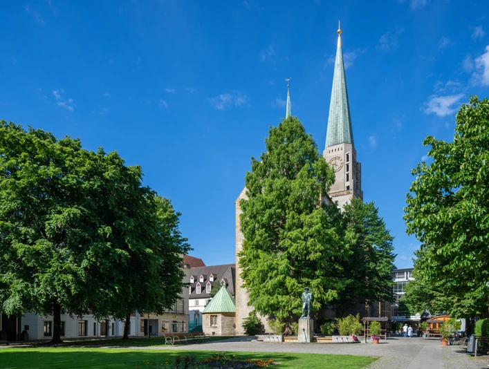 Bielefeld-Kirchplatz-Teutoburger-Wald-Tourismus-Patrick-Gawandtka-001-CC-BY-SA.jpg Die Altstädter Nicolaikirche in Bielefeld, umgeben von Bäumen und einem sonnigen blauen Himmel.