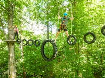 Bielefeld-Kletterpark-Teutoburger-Wald-Tourismus-D-Ketz-039.jpg Ein Mann balanciert auf hängenden Reifen im Hochseilgarten, von Bäumen umgeben.