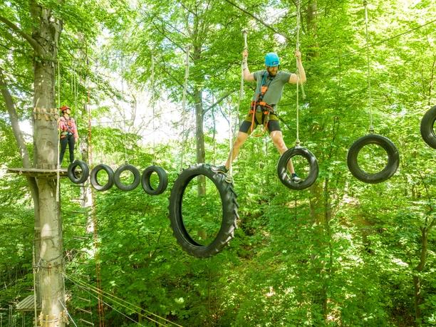 Bielefeld-Kletterpark-Teutoburger-Wald-Tourismus-D-Ketz-039.jpg Ein Mann balanciert auf hängenden Reifen im Hochseilgarten, von Bäumen umgeben.