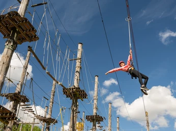 Kletterpark mit hängender Person in Sicherheitsausrüstung vor blauem Himmel und Seilparcours.
