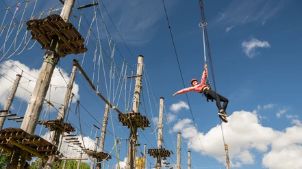 Bielefeld-Kletterpark-Teutoburger-Wald-Tourismus-D-Ketz-028.jpg Kletterpark mit hängender Person in Sicherheitsausrüstung vor blauem Himmel und Seilparcours.