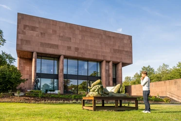 Bielefeld-Kunsthalle-Teutoburger-Wald-Tourismus-D-Ketz-052.jpg Ein Besucher betrachtet eine Skulptur vor einem modernen, kubischen Museumsgebäude bei sonnigem Wetter.