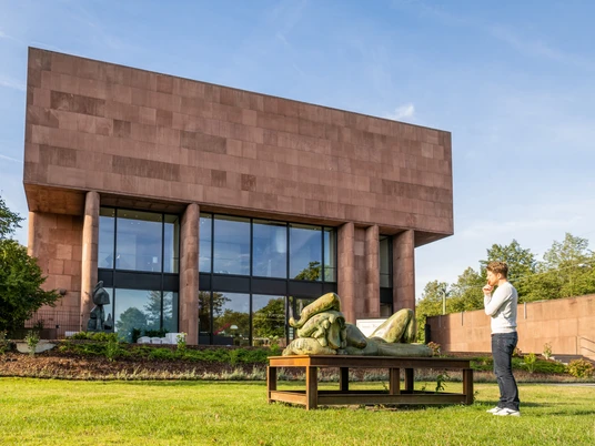 Bielefeld-Kunsthalle-Teutoburger-Wald-Tourismus-D-Ketz-052.jpg Ein Besucher betrachtet eine Skulptur vor einem modernen, kubischen Museumsgebäude bei sonnigem Wetter.