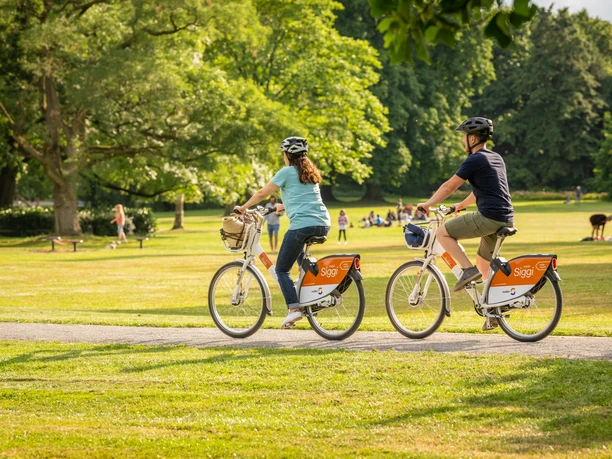 Bielefeld-Nordpark-Teutoburger-Wald-Tourismus-D-Ketz-107.jpg Zwei Menschen fahren mit Fahrrädern durch einen grünen Park, umgeben von Bäumen und entspannten Besuchern.