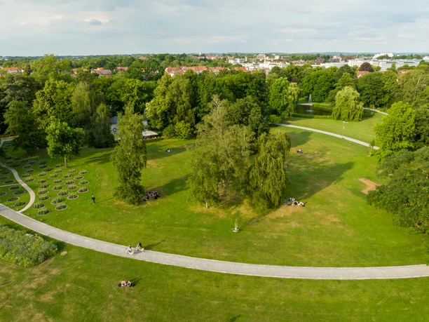 <p>Weitläufiger Park mit Rasenflächen, Bäumen und Wegen, Menschen entspannen im Grünen.</p>
