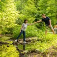 Ein Mann hilft einer Frau, über einen kleinen Bach in einem grünen Wald mit Fahrrädern im Hintergrund.