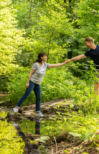 Ein Mann hilft einer Frau, über einen kleinen Bach in einem grünen Wald mit Fahrrädern im Hintergrund.