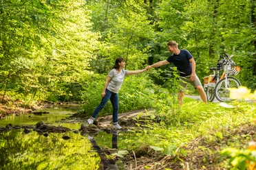 Bielefeld-Oberes Luttertal-Teutoburger-Wald-Tourismus-D-Ketz-088.jpg Ein Mann hilft einer Frau, über einen kleinen Bach in einem grünen Wald mit Fahrrädern im Hintergrund.