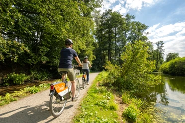 Bielefeld-Oberes Luttertal-Teutoburger-Wald-Tourismus-D-Ketz-083.jpg Zwei Radfahrer auf einem sonnigen Waldweg am Seeufer, umgeben von üppigem Grün und klarem Himmel.