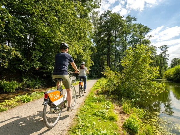 Bielefeld-Oberes Luttertal-Teutoburger-Wald-Tourismus-D-Ketz-083.jpg Zwei Radfahrer auf einem sonnigen Waldweg am Seeufer, umgeben von üppigem Grün und klarem Himmel.