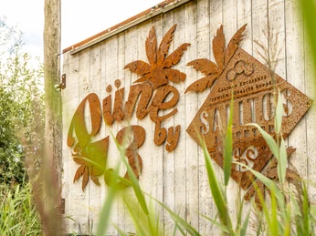 Bielefeld-Obersee-Duene 13-Teutoburger-Wald-Tourismus-D-Ketz-055.jpg Rostige Metallbuchstaben bilden den Schriftzug "Düne by Santo's" auf einer Holzwand mit Gras im Vordergrund.