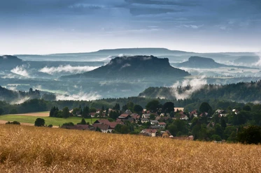 View from the Hohburkersdorfer Rundblick A picturesque landscape with a village in the foreground, surrounded by fields and forests, with misty hills and a cloudy sky in the background.