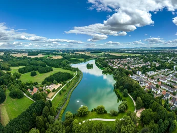 Luftaufnahme eines Sees inmitten grüner Landschaft und Wohngebieten in Bielefeld unter blauem Himmel.