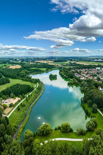 Bielefeld-Obersee-Teutoburger-Wald-Tourismus-D-Ketz-053.jpg Luftaufnahme eines Sees inmitten grüner Landschaft und Wohngebieten in Bielefeld unter blauem Himmel.