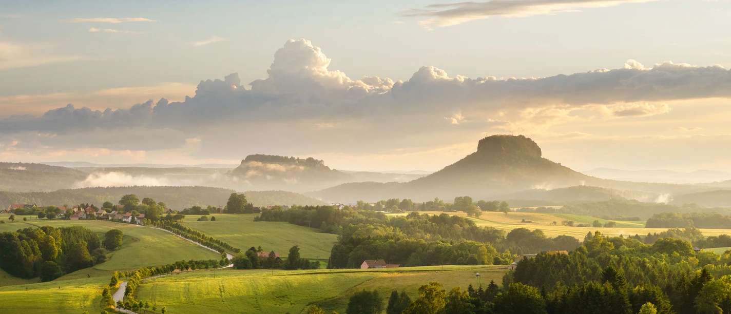 Uitzicht van de Adamsberg naar de Lilienstein.jpg Uitgestrekt groen landschap met glooiende heuvels, een kronkelend pad en twee opvallende tafelbergen op de achtergrond onder een bewolkte hemel.
