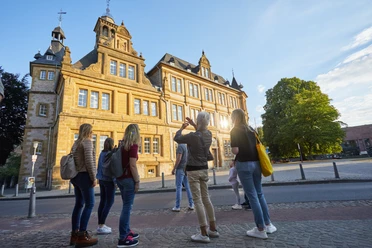 Bielefeld-Ratsgymnasium-Stadtführung-Teutoburger-Wald-Tourismus-T-Evers-092.jpg Gruppe von Touristen besichtigt ein historisches Gebäude in Bielefeld bei Sonnenuntergang.
