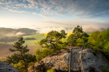 Blick von der Kaiserkrone Felsvorsprung mit Geländer, umgeben von grünen Bäumen, mit Blick auf neblige Felder und Hügel im Morgenlicht.